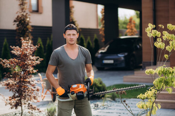 Full length portrait of strong male gardener using electric trimmer for shaping overgrown hedge outdoors. Concept of landscaping and seasonal work