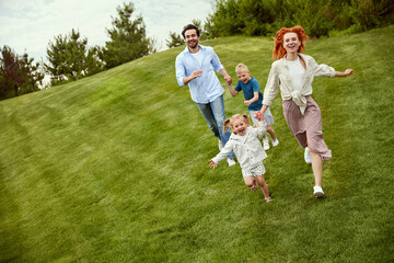 Fototapeta premium Happy couple, man and woman going for a walk in park with their little children, boy and girl, running, playing on grass. Concept of family, childhood and parenthood, fun, weekends, love, ad