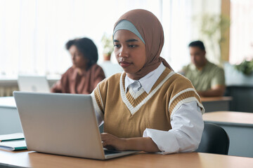 Muslim girl sitting at desk and using laptop during lesson in classroom