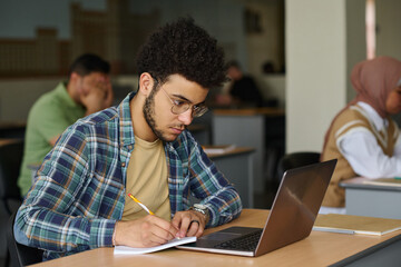 Young man sitting at desk with laptop and making notes in notepad, he learning language in class