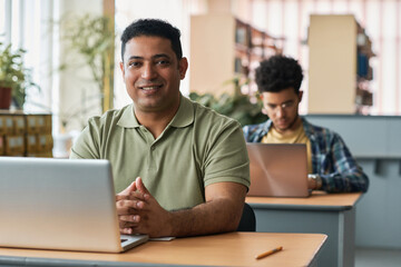Portrait of adult student smiling at camera while sitting at desk with laptop and studying foreign...