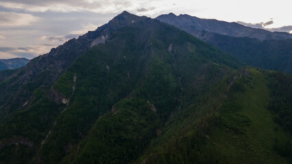 Fototapeta premium Aerial view of the high valley of the rocky mountain range, on which snow lies in places, near to the fast mountain river at summer cloud morning, blue color of the image