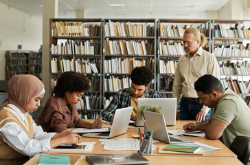 Group of foreign students learning language sitting at table with computers and textbooks together with teacher explaining material