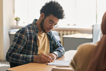 Migrant student sitting at table and writing exam with other students in class