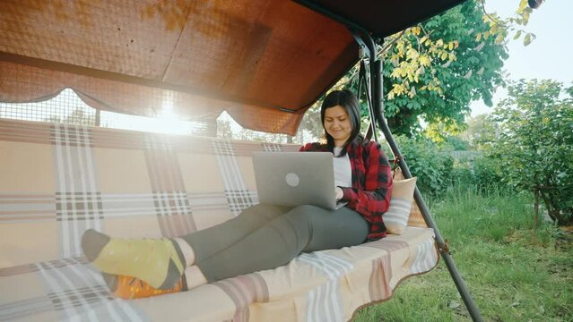 Freelancer girl sits on a swing, working on a laptop, typing text.
