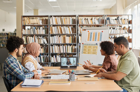 Group of foreign students having online lesson with teacher while sitting in the library