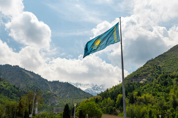 The flag of the Republic of Kazakhstan on top of a mountain, against a background of blue sky with white clouds.