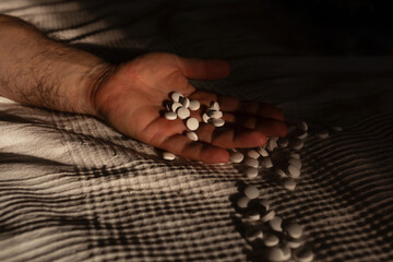 Male hand lying on bed holding bunch of white pills, many spilling onto the bed