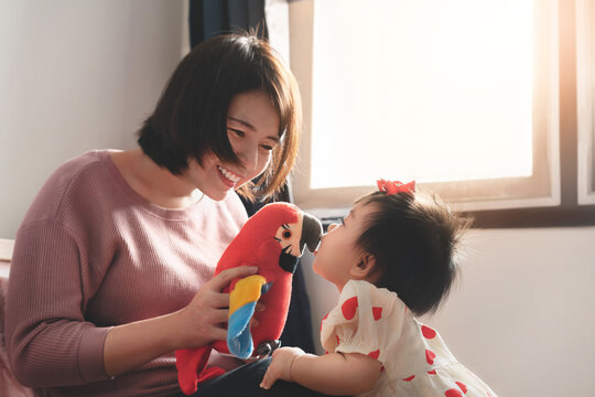 Happy asian woman play parrot doll with her daughter together on bedroom at morning, Baby girl enjoy and kiss doll, Living lifestyle family indoors concept