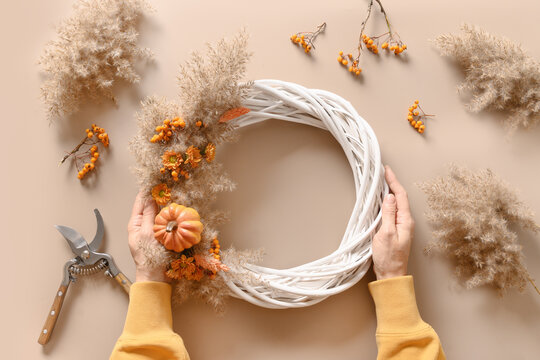 Woman making fall wreath with natural dry flowers and pumpkin on beige table. Top view. Thanksgiving day decoration.