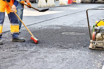 Road workers use a broom and shovel to level freshly laid asphalt around a sewer manhole.
