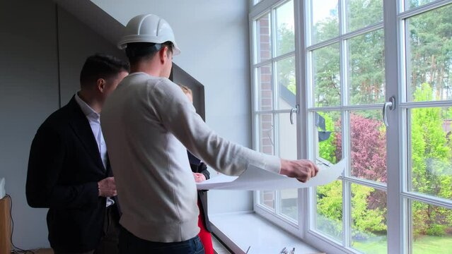 Architect in white helmet talks to wealthy couple about details of renovating summer residence. Builder drawing up plans of suburban cottage