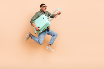 Full length photo of cool hurrying man wear khaki shirt jumping high holding luggage empty space isolated beige color background