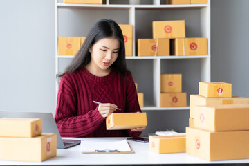 Female business owner holding parcel box for delivery and using smartphone to sell online at home office. SME business operators