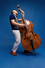 Full-length portrait of young man, musician, emotionally, excessively playing double bass against blue studio background. Concept of music, talent, hobby, entertainment, festival, performance, ad - obrazy, fototapety, plakaty