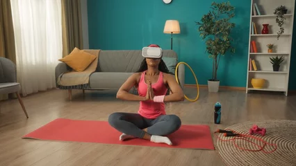 Fotobehang African American woman in virtual reality glasses sits in a lotus position. A young woman in a pink tracksuit is doing yoga in virtual reality. Lifestyle and technology concept. © kinomaster