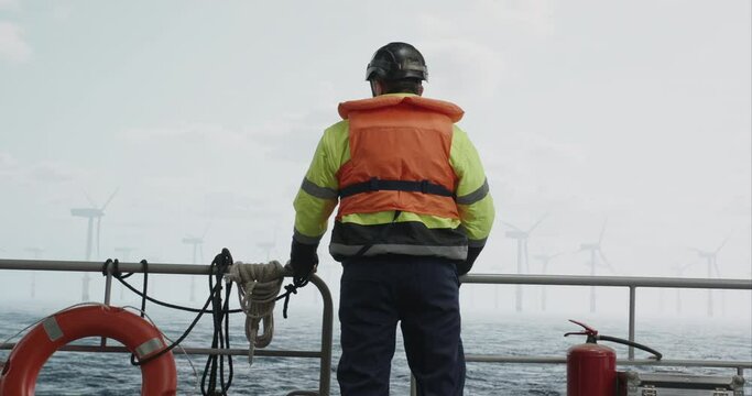 Maintenance engineer standing on a service vessel approaching offshore wind turbine farm. Portrait of skilled professional preparing to work at high altitude