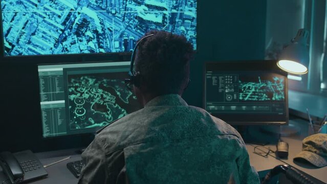 Back view of male African American military surveillance officer sitting in wheelchair at desk in front of computer screens monitoring situation and reporting during call in headset