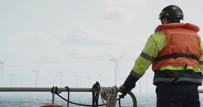 Maintenance engineer standing on a service vessel approaching offshore wind turbine farm. Portrait of skilled professional preparing to work at high altitude