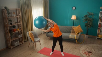 Elderly woman goes in for sports with a fitness ball in the living room. A greyhaired woman holds a ball over her head and does side bends. View from above. Keeping body in tone.