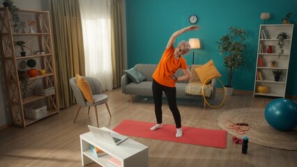 Elderly woman exercising in front of a laptop in the living room. A woman is watching a video tutorial on a laptop. A grayhaired retired woman makes side bends. Keeping the body in good shape.