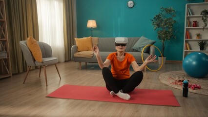 A retired woman in virtual reality glasses sits in a lotus position. Elderly woman doing yoga in virtual reality. Yoga class and technology.