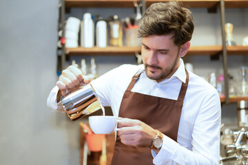 Handsome Caucasian waitress small business owner barista a making latte or cappuccino coffee pouring milk making latte art at the cafe. Man working with small business owner or SME concept. - obrazy, fototapety, plakaty