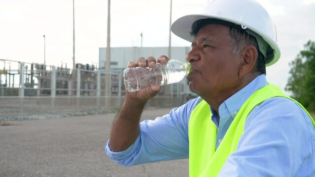 Elderly Asian male worker or engineer drinking refreshing water on summer day near wind turbine power plant.