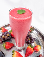 A glass glass with juice with red, dark pink berries, grapes and strawberries on a tray.