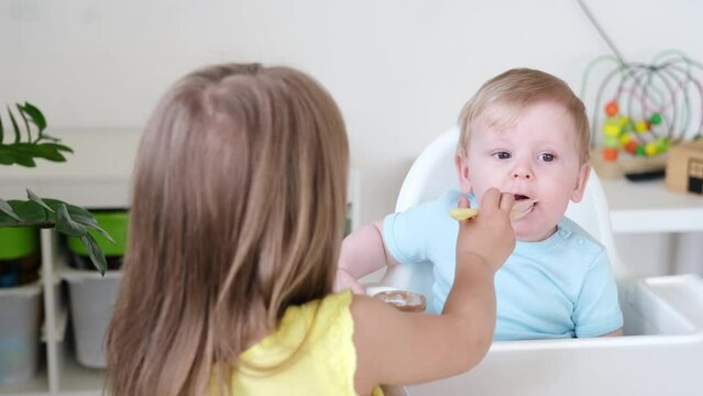 baby boy eating bland mashed food sitting on high chair, older sister feeding child 