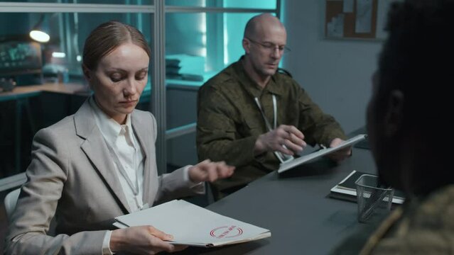 Tilt shot of male African American military officer giving secret documents to serious Caucasian woman and man during official briefing indoor at night