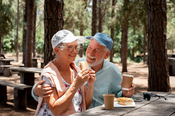 Smiling senior family couple enjoying a sandwich while sitting at a wooden table in the woods appreciating nature and freedom, happy retired seniors man and woman and healthy lifestyle concept