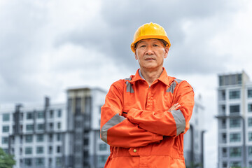 Serious and determined middle age Asian engineer in orange jumpsuit with hard hat crossing his arms while standing proudly in front of his multi-stories buildings