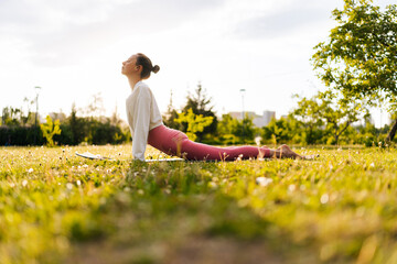 Low-angle side view of sportive and healthy young woman in white sportswear practicing yoga in green park doing Upward facing dog yoga pose on sporty mat. Concept of outdoors yoga, fitness and active.