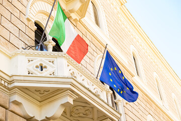 The Italian and European flags are waving on a balcony of a Baroque-style institutional building.