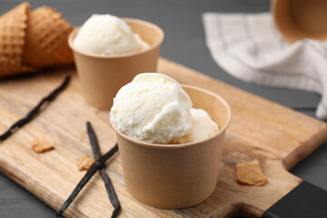 Paper cups with delicious ice cream and vanilla pods on wooden board, closeup