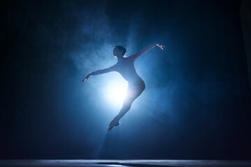 Expressive, artistic young woman, professional ballerina making creative performance against dark blue background with spotlight