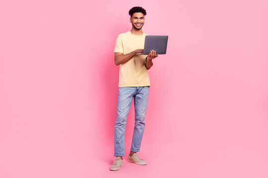 Full length photo of clever smart cheerful guy wear yellow shirt denim pants hold laptop texting letter isolated on pink color background