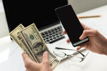 Silhouette of cropped shot of a young man working from home using smart phone and notebook computer, man's hands using smart phone in interior, man at his workplace using technology, flare light