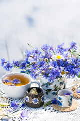 Obraz na płótnie Canvas Cornflower herbal tea in white cup on white crochet napkin on wooden table outdoors, picnic with healthy cornflower drink with honey in nature background, healthcare and healthy eating concept