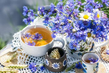 Cornflower herbal tea in white cup on white crochet napkin on wooden table outdoors, picnic with healthy cornflower drink with honey in nature background, healthcare and healthy eating concept