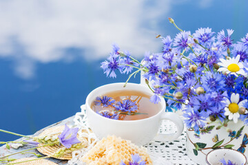 Cornflower herbal tea in white cup on white crochet napkin on wooden table outdoors, picnic with healthy cornflower drink with honey in nature background, healthcare and healthy eating concept