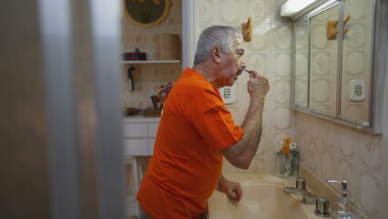 Candid senior man shaving beard in front of mirror in bathroom. Authentic real life old age...