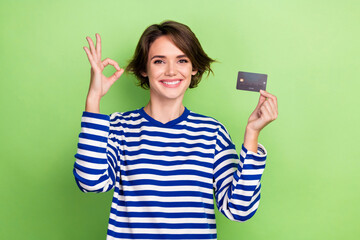 Portrait of optimistic positive woman wear striped sweatshirt hands hold card showing okey approve isolated on green color background