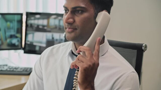 Medium close up portrait of Indian security manager talking on phone while sitting at desk in office and looking at camera