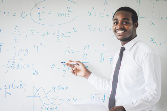 Portrait of smiling african teacher in white shirt standing near blackboard with formulas on it.Concept of sciences education