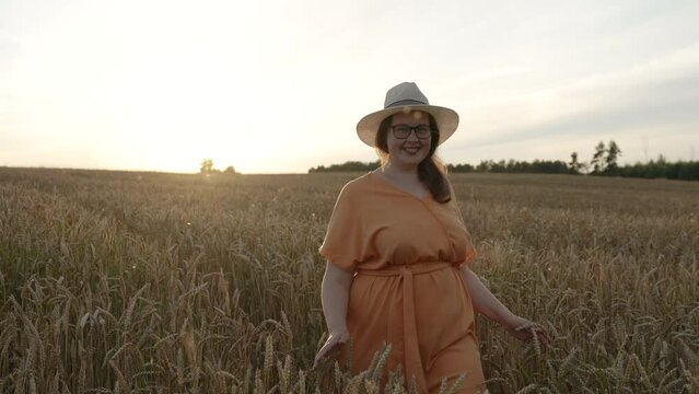 Attractive plus-size middle-aged woman with glasses smiles, walks alone through cereal field. Lady in hat, orange dress is touching spikes with hands. Concept of self-acceptance, self-love, myopia.