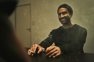 Man, suspect smoking and detective in interrogation room with criminal, terrorist or interview with a gangster. Police officer, murder questioning and statement on crime and person with cigarette