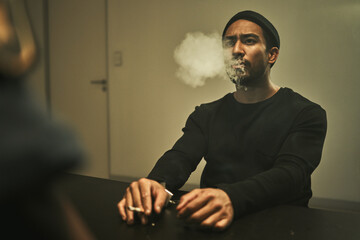 Man in handcuffs, smoking and talking to detective in interrogation room with criminal, suspect or...