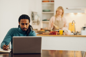 An Arabic freelancer is sitting at home and working on a project on a laptop at the dining table while his spouse is cooking dinner.